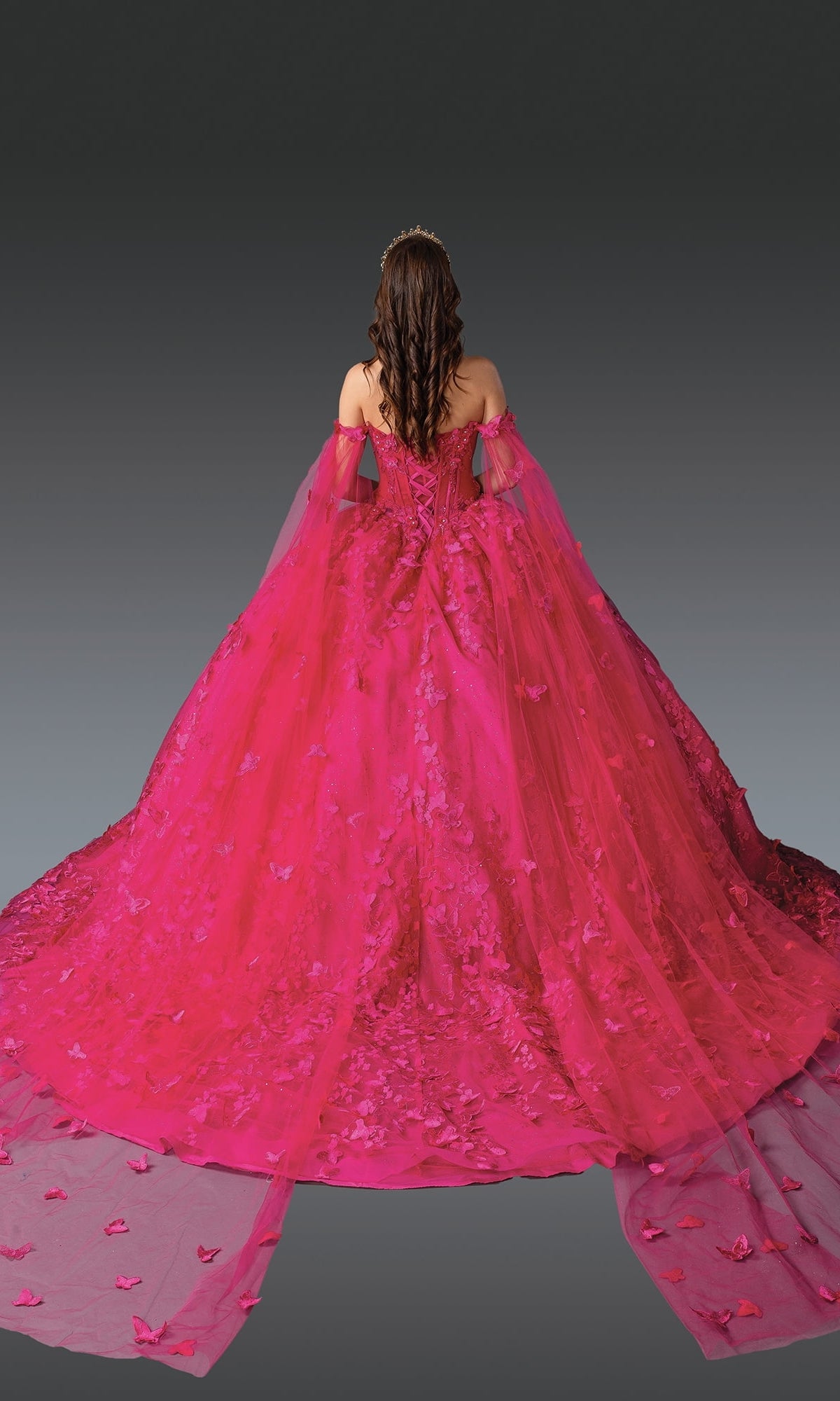 A woman with long wavy hair wears the Fuchsia Quinceanera Dress 1960 By Dancing Queen, featuring a corset bodice, floral details, and butterfly embroidery, standing against a gray gradient background.